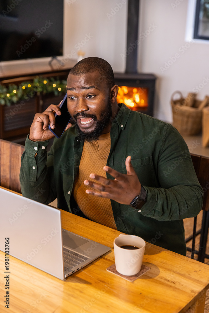 Obraz premium African American man talking on phone, gesturing, working at laptop in home office with coffee mug