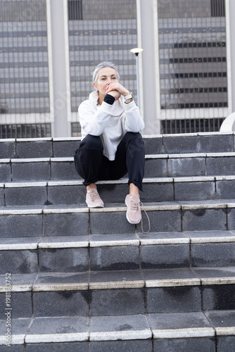 Senior woman sitting on stone steps in urban plaza, wearing hoodie, resting and gazing after rain
