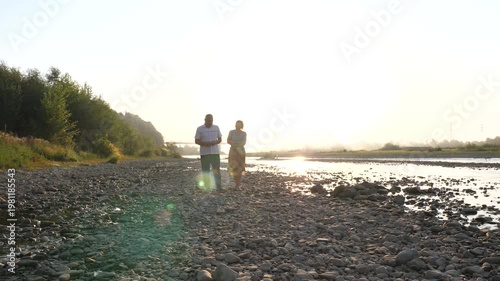 A man and a woman stand on a rocky riverbank watching the water under soft evening light