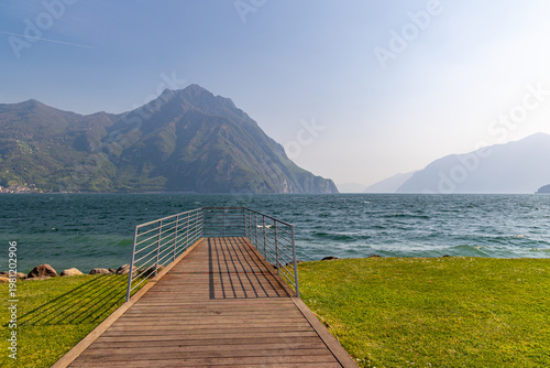 Wooden pier overlooking the scenic landscape of Lake Iseo, Italy, with majestic mountains and calm blue waters under a clear sky.