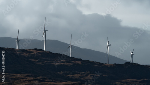 tall white wind turbines stand on a rugged brown hill, with hazy mountains in the background under a cloudy sky.