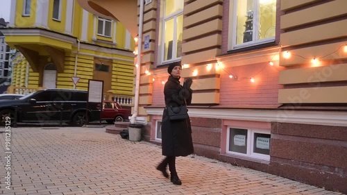 Elegant Woman in Black Winter Coat Walking on Illuminated City Street at Dusk with Festive String Lights