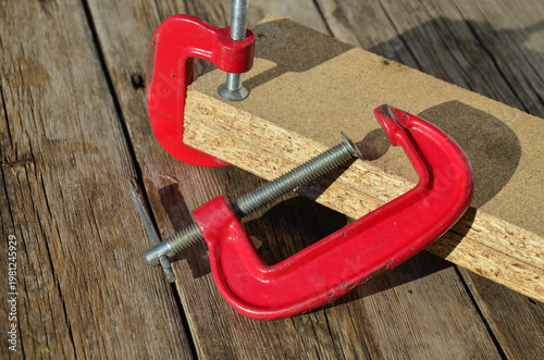 Red C-clamp on a wooden workbench background.
