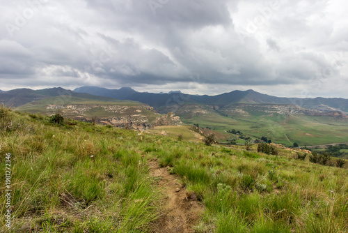 A small hiking trail high up in the Drakensberg Mountains of South Africa, with storm clouds gathering over the distant blue peaks in the background.