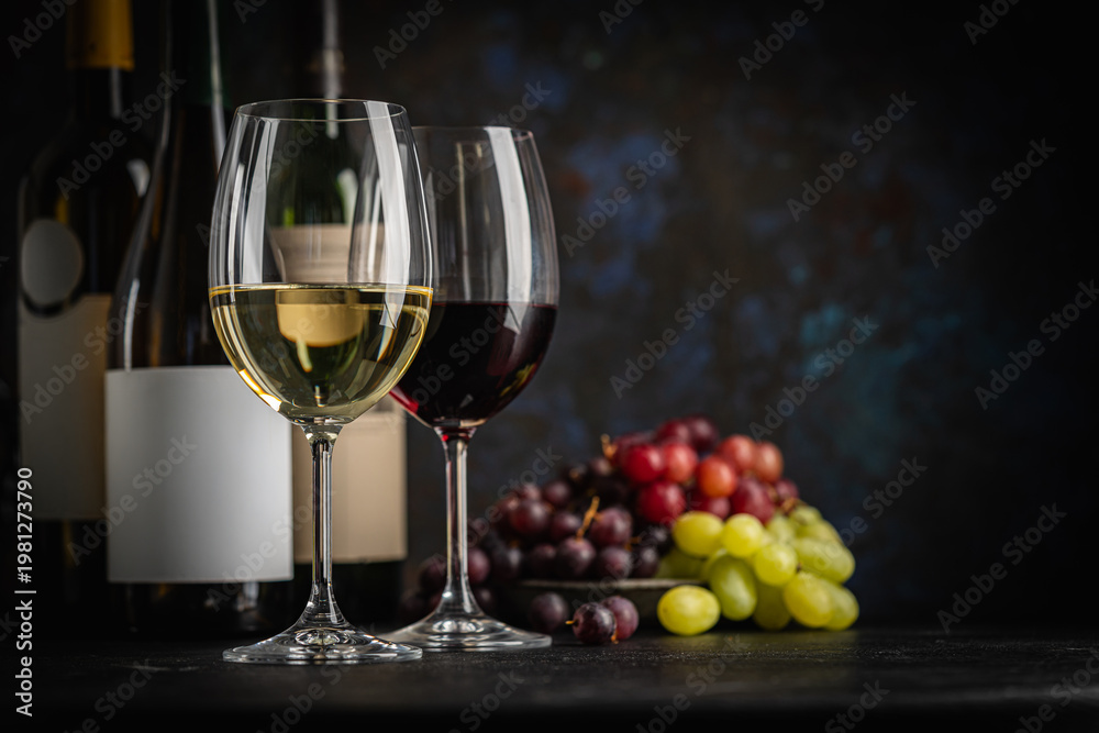 Naklejka premium A dark, elegant still life of White and Red wine in a glass with wine bottles and fresh grapes in the background. Perfect for wine-themed compositions