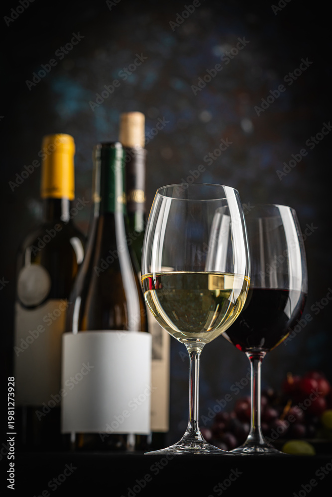 Fototapeta premium A dark, elegant still life of White and Red wine in a glass with wine bottles and fresh grapes in the background. Perfect for wine-themed compositions