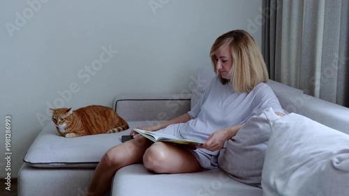 Woman is sitting on a sofa with her ginger cat, trying to decide whether to spend time using her smartphone or reading her book