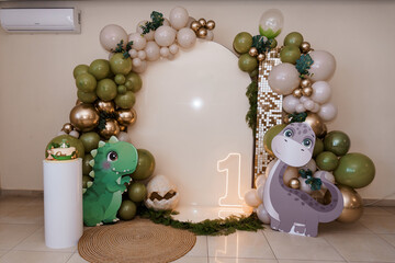 First birthday dinosaur party balloon arch photo backdrop with green and gold decorations and cute cartoon dino cutouts