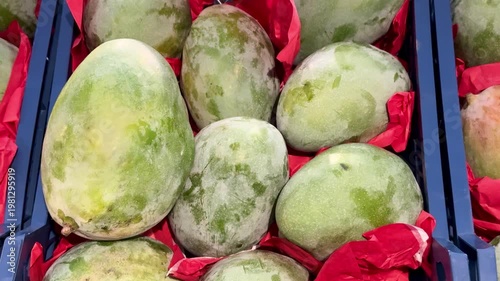 Green Mangoes In Plastic Crate Overflowing At Bustling Fruit Stall, Glossy Skins Resting On Red Tissue, Blue Crate Edges Visible.