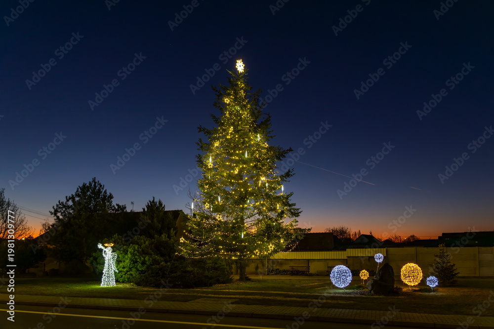 Fototapeta premium Christmas tree and festive decorations lighting up Chvalovice at dusk