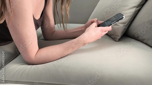 Woman lies on sofa and types message on smartphone at home. Female writes text and replies using mobile device in messenger. Concept communication, chatting, online interaction, digital