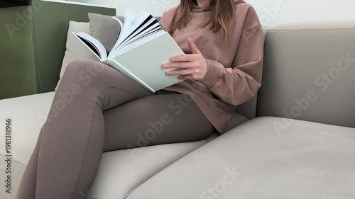 Woman quickly flips through book while sitting on sofa at home. Female scans pages fast reviewing content indoors. Information search and reading overview concept in apartment living room..