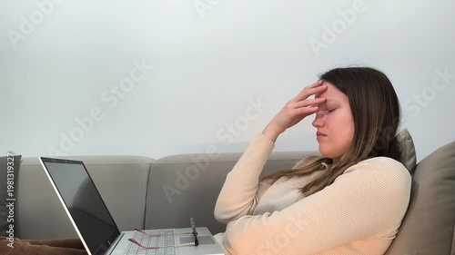 Laptop work pauses as woman on sofa holds head with closed eyes from migraine. Female experiences headache during remote job using computer at home. Health concept related to stress and