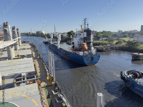 Schilderij op canvas Large blue cargo ship moving through narrow industrial canal with tugboat assist