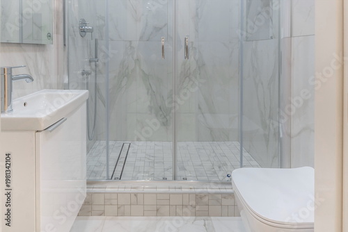 Quadro su tela A bright bathroom decorated with light marble tiles, glass shower doors and a white sink and toilet