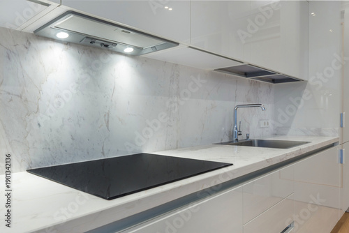 A fragment of a white kitchen set with a light countertop, sink, built-in electric stove panel and extractor hood.