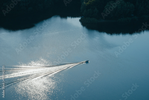 Aerial view of small motorboat cruising on calm blue lake water with sun reflection. Drone shot of boat leaving white wake trail at sunset evening time.