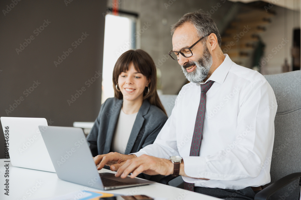 Fototapeta premium A senior professional man and a young female colleague working side-by-side on their laptops in a modern office