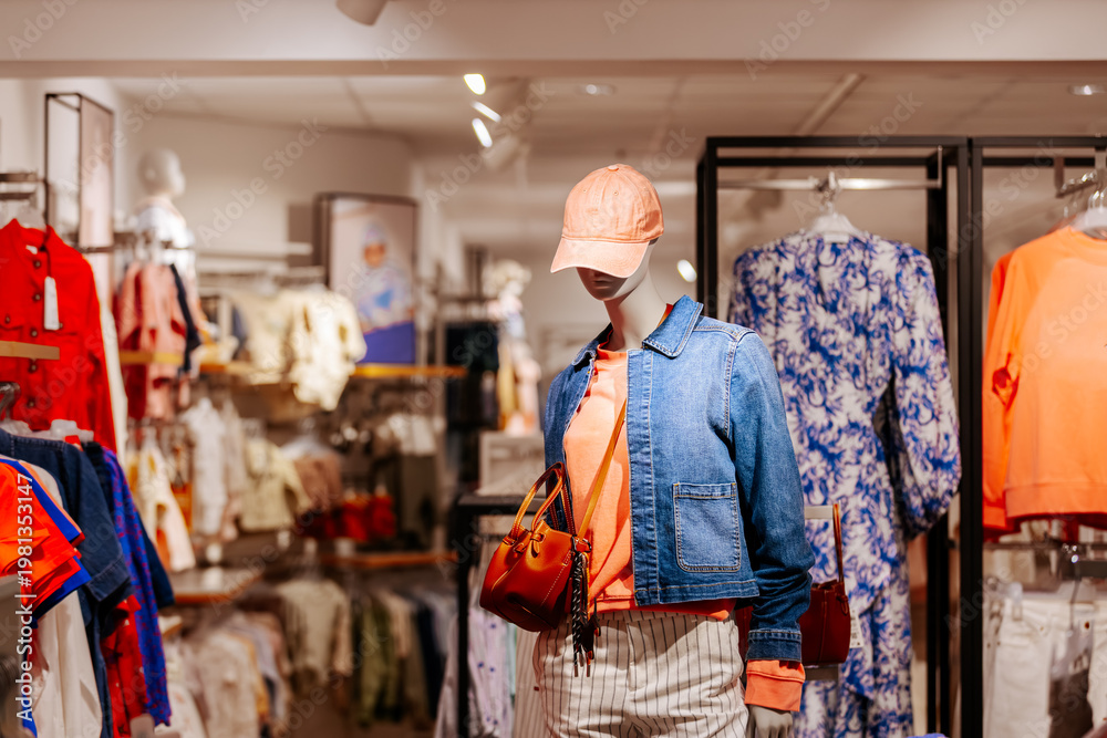 Obraz premium Female mannequin dressed in denim jacket, orange sweater, and striped pants stands in a clothing store with various garments displayed on racks in the background