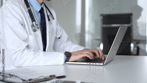 Foto Confident doctor wearing a white coat and stethoscope, using a laptop at a desk for medical record keeping and online patient care