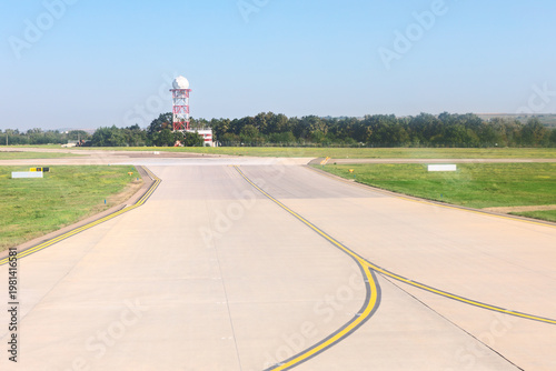 Wide airport taxiway stretches forward under a clear sky, bordered by green grass and trees. In the distance, a red and white radar tower stands near the horizon