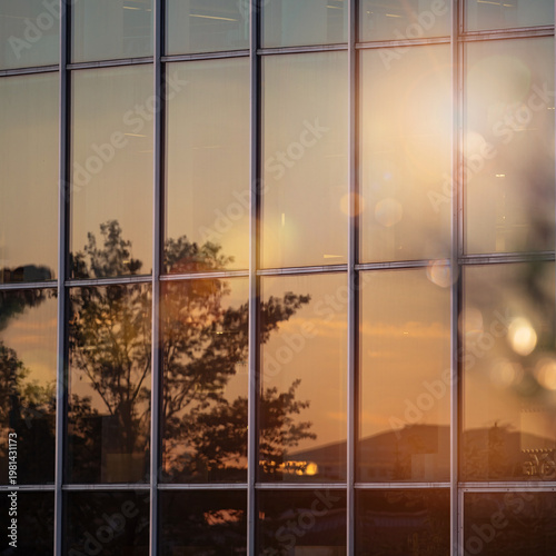 Large grid patterned glass and metal office building facade reflecting urban landscape and warm sunset with lens flare