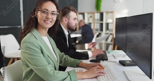Analyst woman in office at computer. She smiles to camera as colleague reads charts, analyzes data and statistics for finance and report insight on dual screens. Concept of teamwork and analytics.