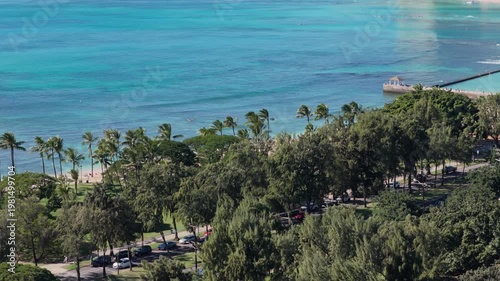 Aerial above city park to reveal Waikiki Beach with palm trees and tall buildings in the background, Honolulu 