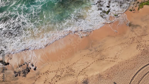 Aerial top down view of a beach with waves crashing onto the shore. Green water and white foam indicating turbulent waves. Warm color sand with footprints