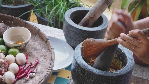 Authentic Thai cooking scene with fresh ingredients and traditional stone mortar and pestle. Hands preparing spicy sauce using chilies, garlic, lime, eggs, and herbs on a rustic kitchen table. Concept