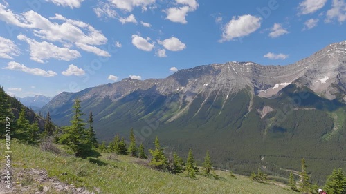 Scenic summer mountain landscape with dramatic rocky peaks, green forest valleys, and blue sky with clouds. View from a hiking trail overlooking the Canadian Rockies, capturing nature, adventure, and 
