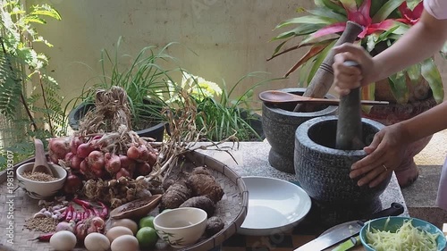 Authentic Thai cooking scene with fresh ingredients and traditional stone mortar and pestle. Hands preparing spicy sauce using chilies, garlic, lime, eggs, and herbs on a rustic kitchen table. Concept