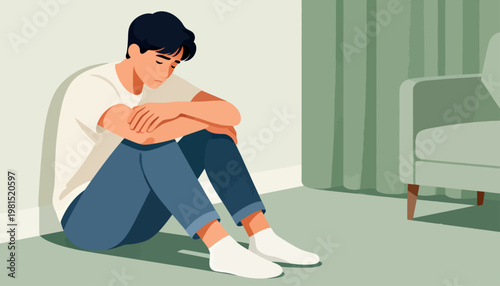 Young man sits hunched against wall, head down. Blue jeans, white t-shirt, and socks complete his casual look. He appears withdrawn, possibly stressed or melancholic