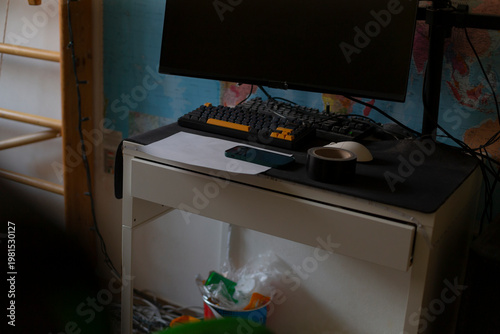 Busy study desk interior with keyboard, scattered wrappers and bag under desk, monitor glowing against map wallpaper, latenight study vibe and casual organization.