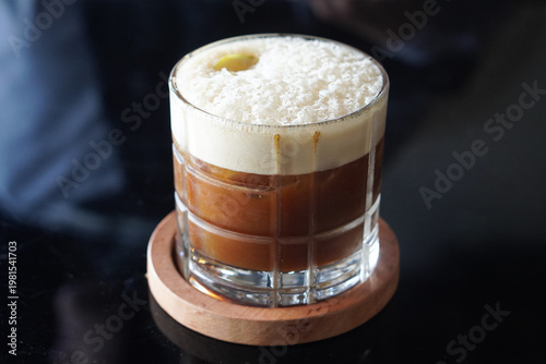 Konstfotografi Creamy signature coffee in a clear glass with thick foam and rich brown tones in a cozy café setting