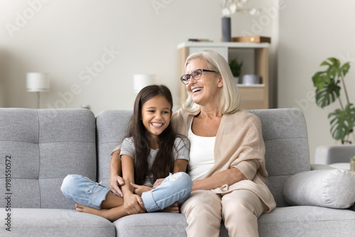 Cute school-age girl spend time with older granny, multigenerational family siting on couch, laughing, lead pleasant, trustworthy conversation, share news, joking, enjoy carefree rest at home together