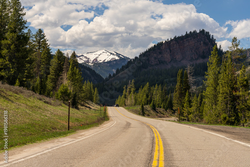 Scenic open highway winding through a lush mountain canyon with snow-capped peaks and pine forests in Wyoming, USA.