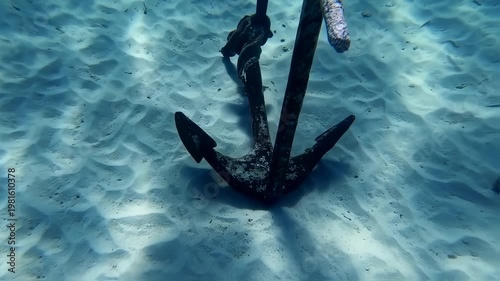 Sunken anchor resting on the sandy seabed in clear blue ocean waters, underwater scene