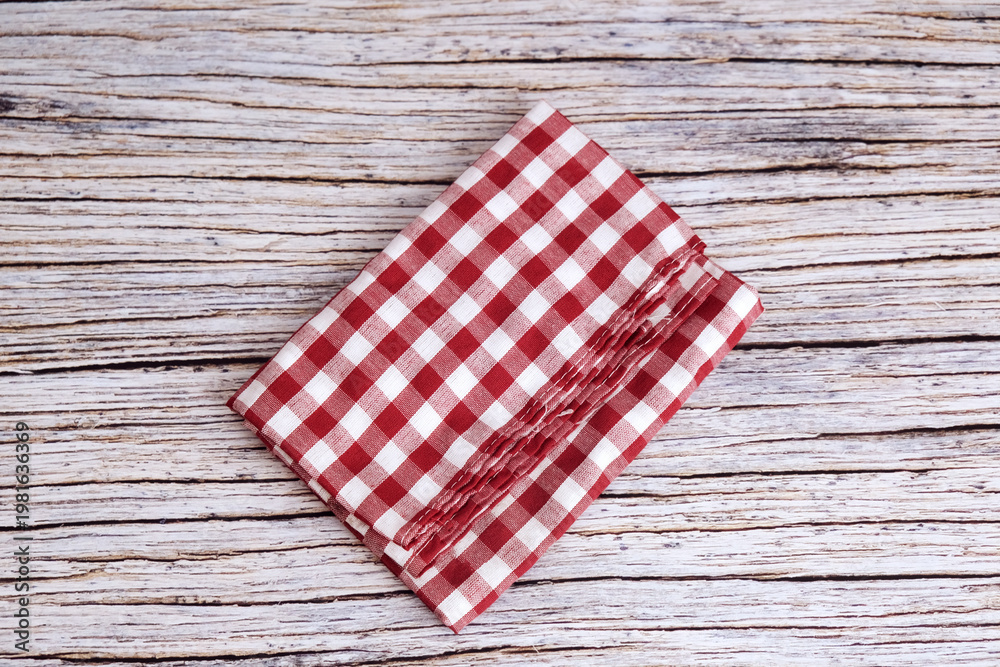 Obraz premium Red and white checkered cloth napkin rests on wood. Folds show soft texture and gentle creases. Rustic background adds charm and nostalgia. Ideal for kitchens, cafes, or picnic scenes