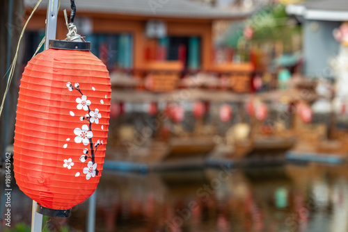 Konstfotografi Vibrant red Japanese lantern adorned with delicate cherry blossoms, hanging against a softly blurred background of traditional buildings and their reflections on water, evoking serene cultural charm