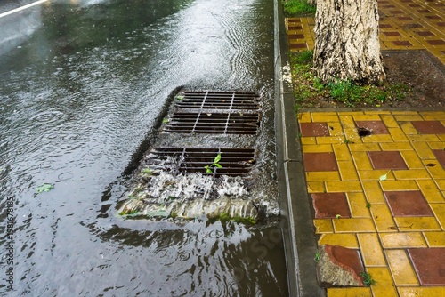 Stormwater rushing into a metal street grate beside a sidewalk with a tree during rainfall in a quiet city scene.