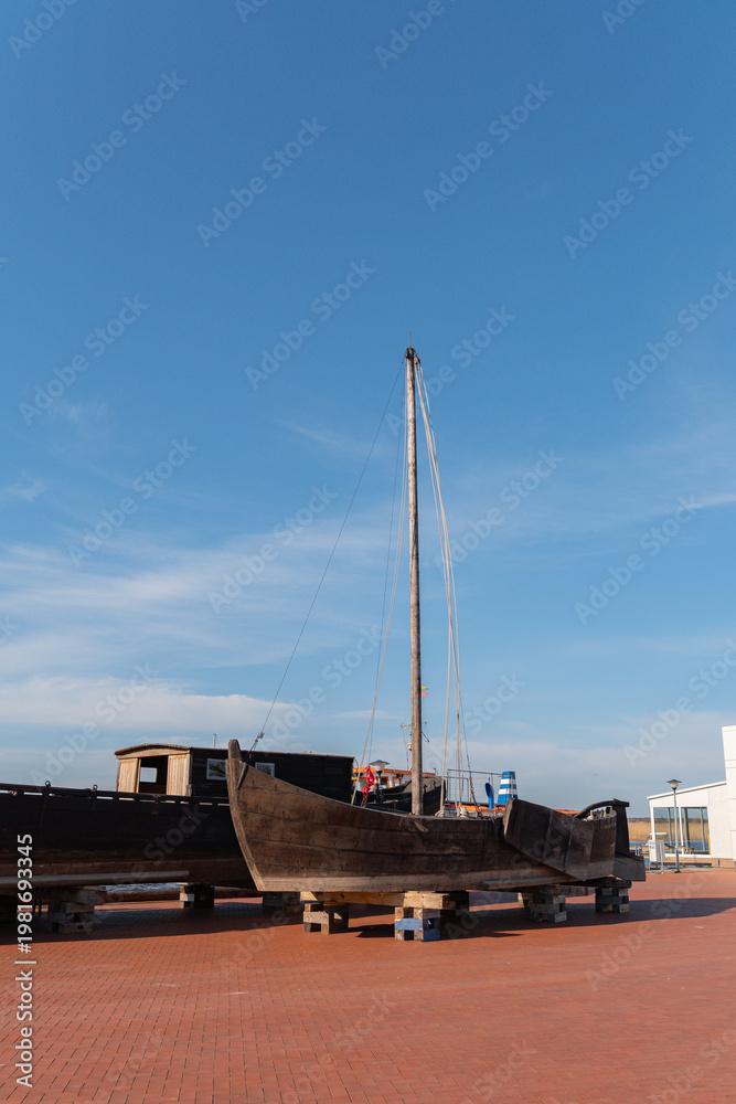 Fototapeta premium Old wooden sailing boat rests on supports outside on a paved pier area under a clear blue summer sky waiting for restoration work to begin in the coming days