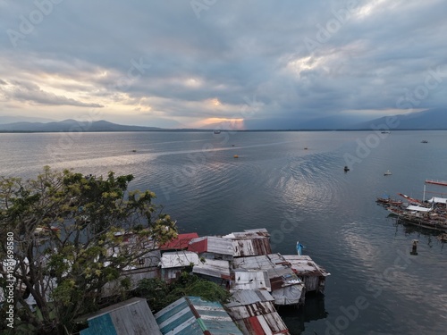 Aerial photograph of weathered corrugated rooftops of a coastal fisherman village at dusk with calm tropical bay mountain silhouette and dramatic sunset sky near Puerto Princesa Palawan