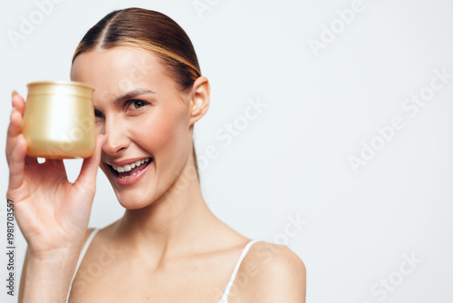 Cheerful woman with a gold cup held near her lips, bright smile, studio portrait. Beauty wellness spa relaxation concept with clean skincare and minimal soft white background.