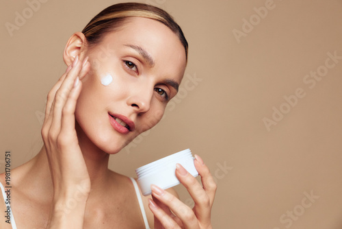 Woman applying face cream in a close up on a beige background, holding a white jar, soft studio lighting, skincare routine, wellness and self care, fresh healthy glow