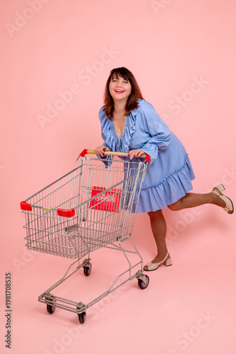 Happy brunette woman 40-50 years old in a blue dress with a shopping cart on a pink background