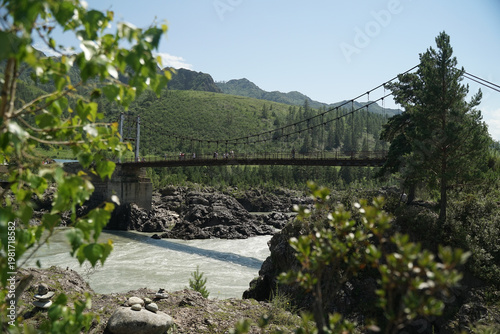 A view of a bridge spanning a rocky riverbed with a strong current, surrounded by lush green trees and mountains