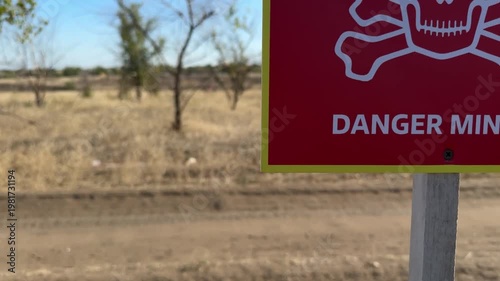 A lone cyclist moves through a dry rural field marked ukrainean words with a red “Danger Mines” warning sign. The scene highlights the risks of unexploded ordnance in post-conflict areas