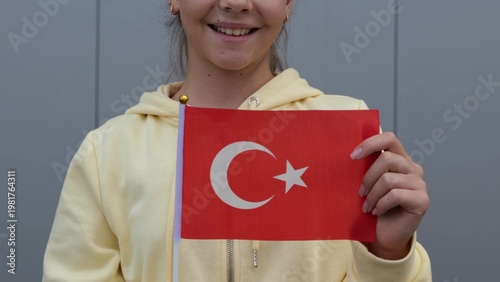 Caucasian teenage Girl waving small flag of Turkey. Zoom out, close up, grey background, female small girl in yellow hoodie, keeping flag of Turkey in her hands.