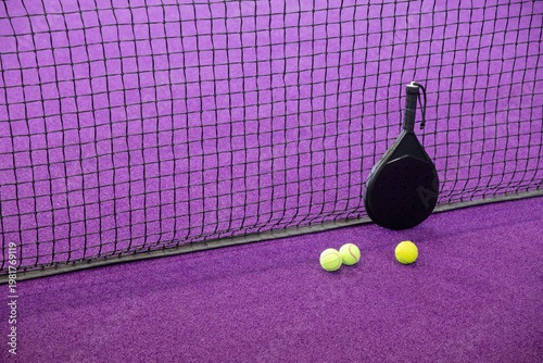 Black padel racket leaning against mesh net on purple turf with three fluorescent balls, copy space